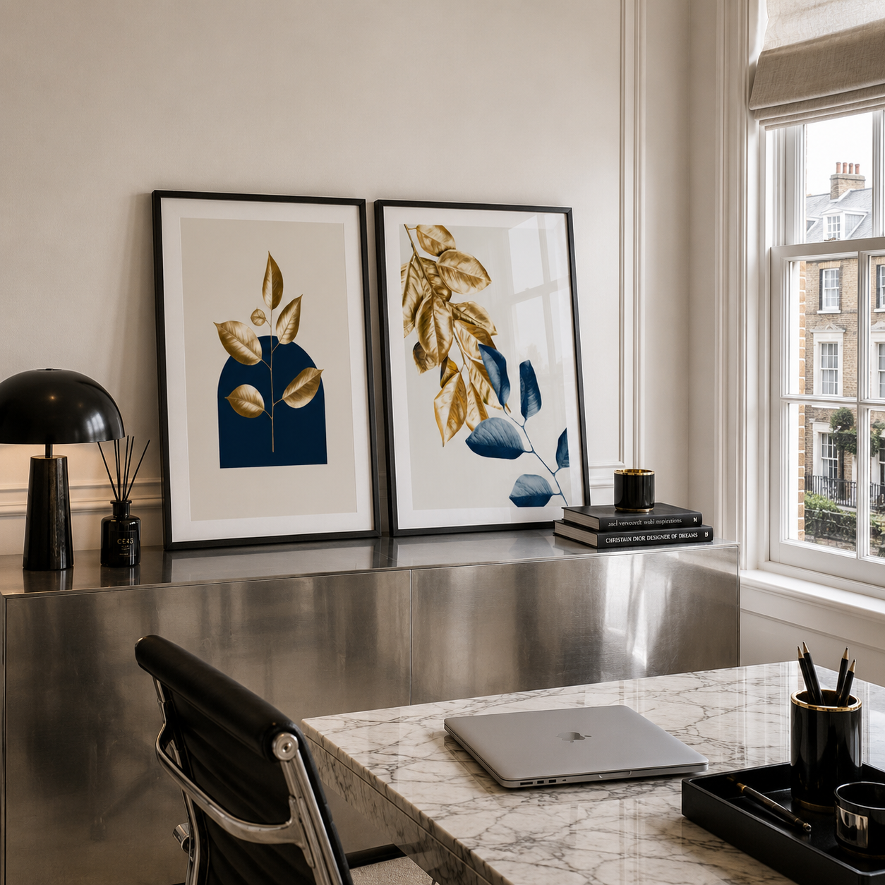 Two framed botanical art prints with gold and blue leaf designs hang on a wall above a metallic desk in a modern home office setting.