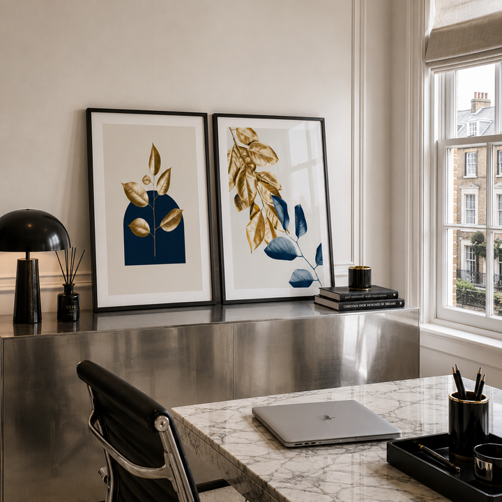 Two framed botanical art prints with gold and blue leaf designs hang on a wall above a metallic desk in a modern home office setting.