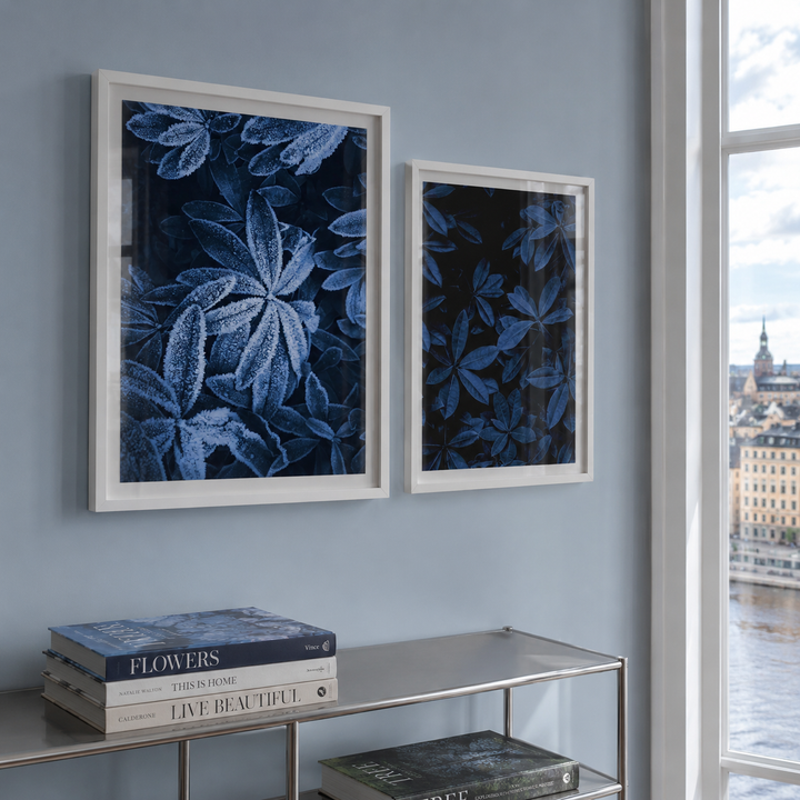 Two framed botanical art prints with deep blue leaf patterns on a dark background, hung on a light blue-gray wall above a glass-top console table. A window on the right shows a river and cityscape.