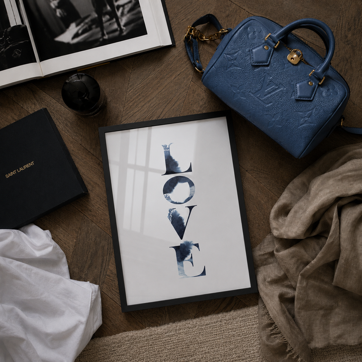 A framed vertical art print featuring the word 'LOVE' in dark blue watercolor-style lettering on a white background, styled in a flat lay with a blue embossed Louis Vuitton Speedy Bandoulière 20 Bag bag, a black Saint Laurent box, an open book, and fabric on a wooden floor.
