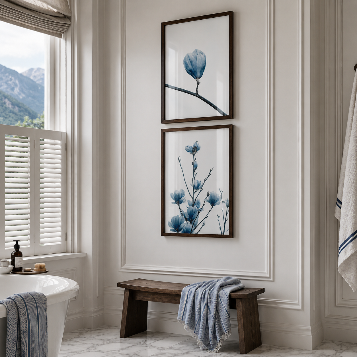 Two vertical framed botanical art prints with blue flowers and branches hang on a white paneled wall above a rustic wooden bench in a bright bathroom setting.