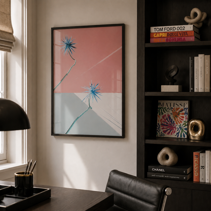 A framed abstract art print hangs on a beige wall in a modern home office setting, featuring a pink and light gray background with blue starburst flower designs. The room includes a dark wood desk, black leather chair, and a dark wood shelving unit with books and decor.