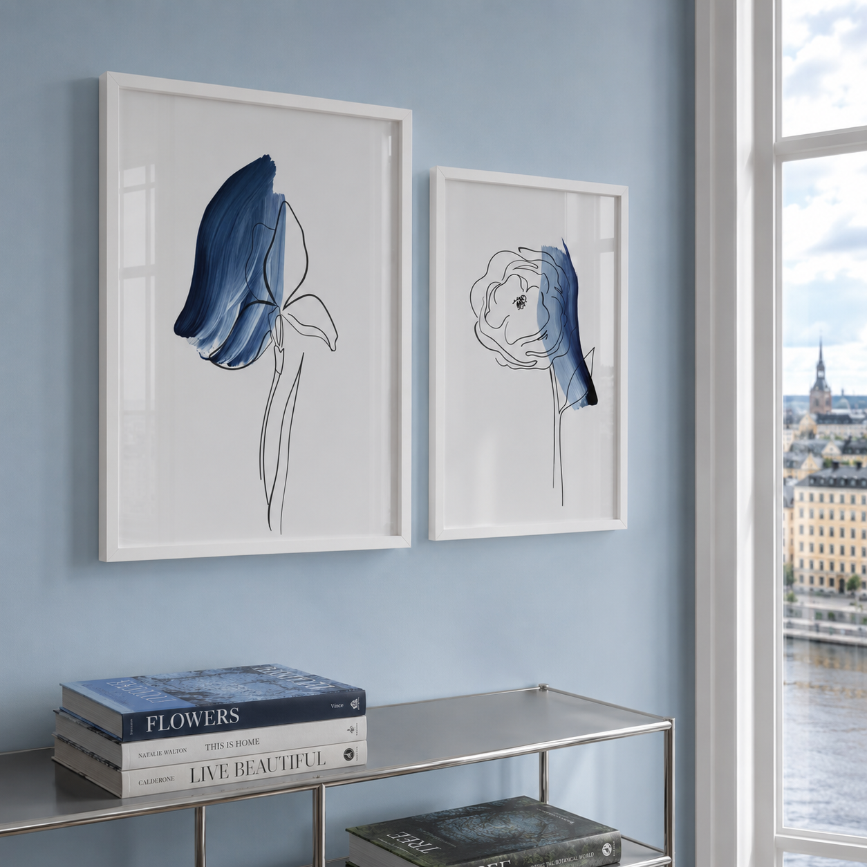 Two framed minimalist floral art prints with blue and black line drawings on white backgrounds, hung on a light blue wall above a glass shelf in a modern room.