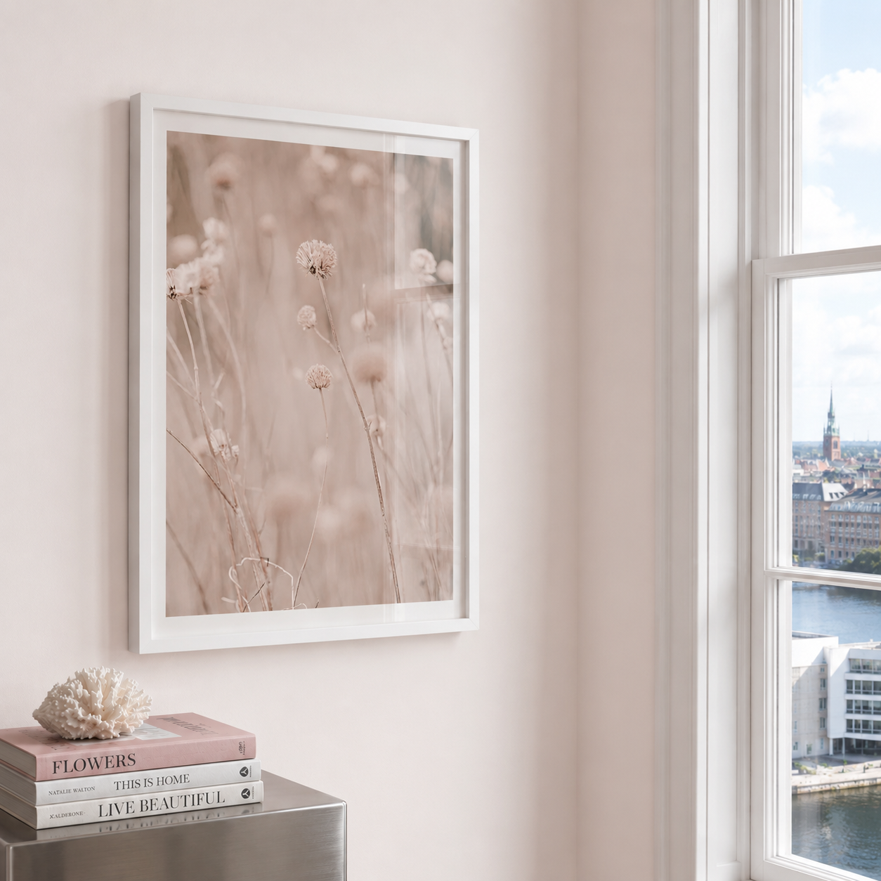 A framed botanical art print featuring soft beige wildflowers on a light wall, styled in a room next to a window with a city view. The frame is white, and below it is a small metallic side table with stacked books and a coral shell.