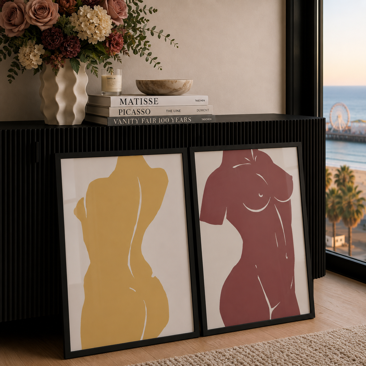 Two framed minimalist Matisse inspired, art prints leaning against a black ribbed cabinet in a styled interior. The left print shows a yellow abstract female torso silhouette, and the right print shows a muted red abstract female torso silhouette.