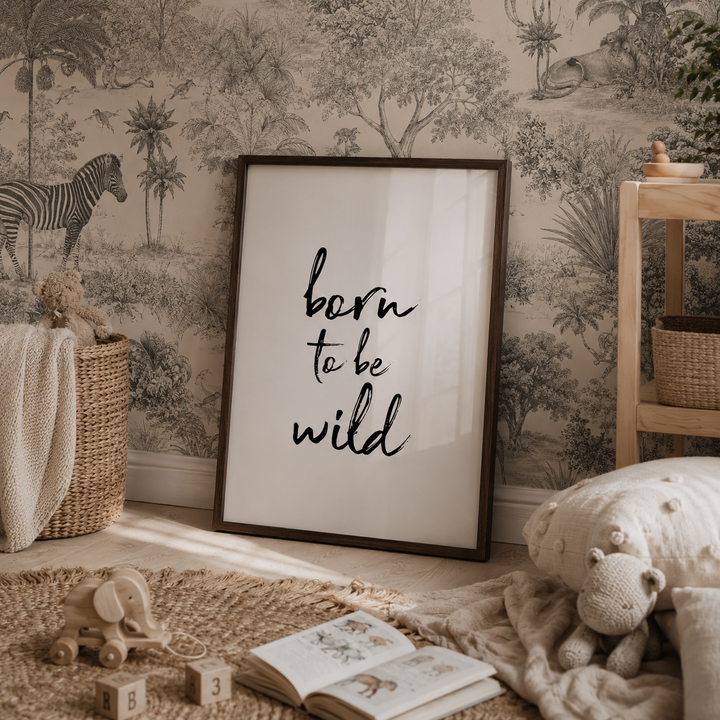A framed wall art print with the phrase 'born to be wild' in black script on a white background, leaning against a safari-themed wallpapered wall in a styled nursery setting with toys, blankets, and a wooden shelf.