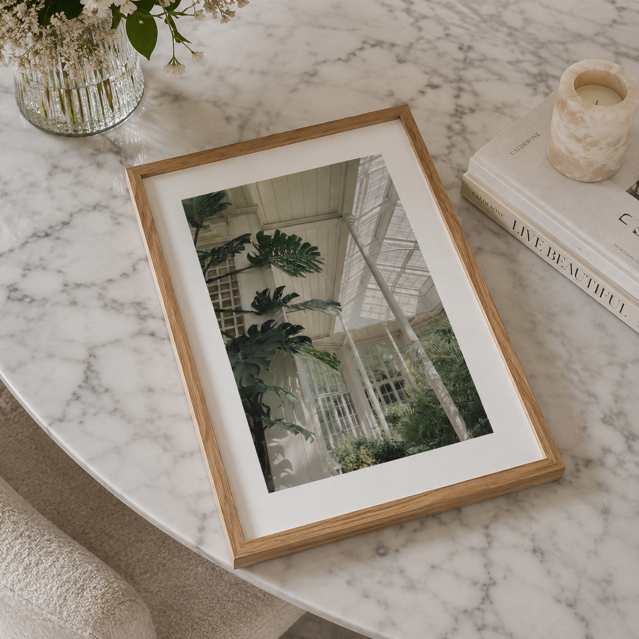 A framed botanical art print of a greenhouse interior, styled on a white marble table next to a glass vase of flowers and a book.