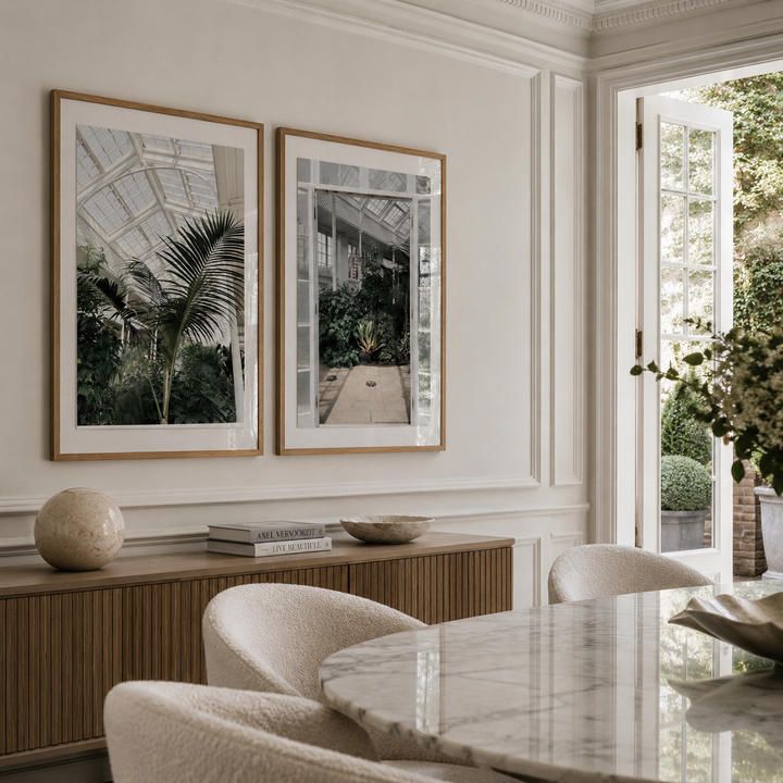 Two framed botanical art prints hang on a cream wall above a wooden console table in a dining room setting. The prints feature black and white architectural greenhouse scenes with green palm leaves, enclosed in light wood frames.