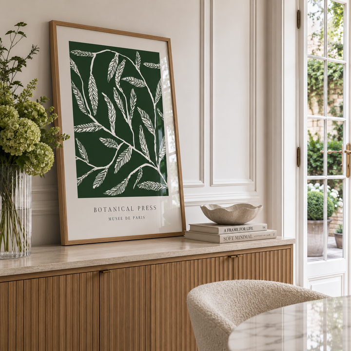 A framed botanical art print with white leaf illustrations on a dark green background, leaning on a light wood credenza in a bright, modern room.