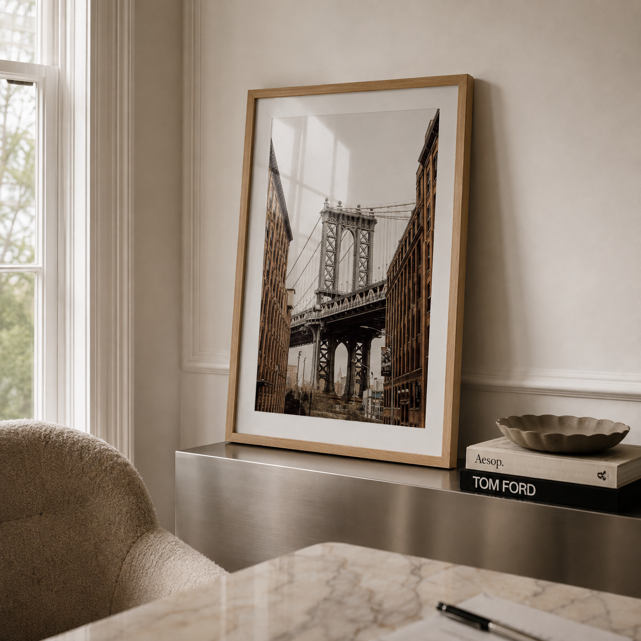 A framed art print of the Manhattan Bridge in New York City, styled in a light wood frame leaning against a cream wall on a metallic console table next to a window.