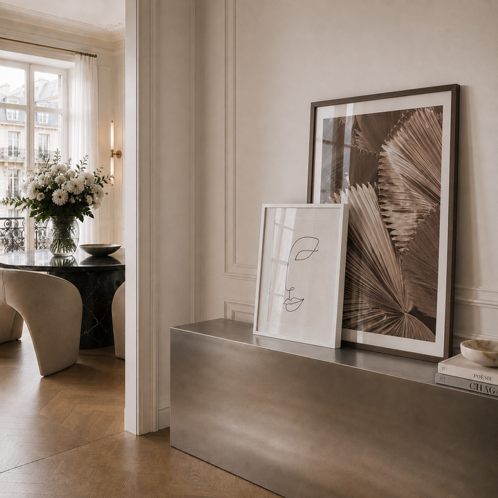 Two framed art prints displayed on a stainless steel console table in a modern, elegant interior. The larger print features a brown palm frond pattern in a dark frame, and the smaller print has minimalist black line art on white paper in a light frame.