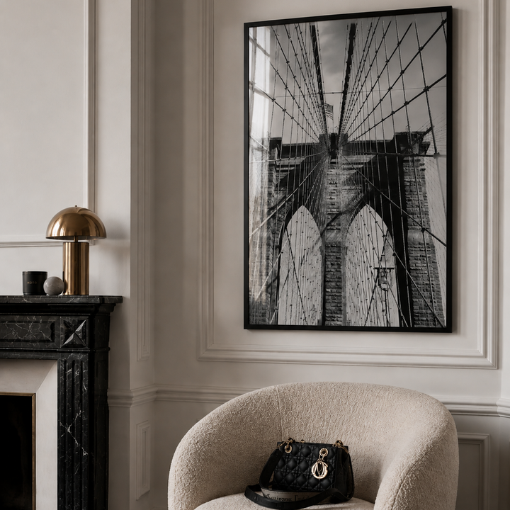 A large black and white framed wall art print of the Brooklyn Bridge hangs on a white paneled wall in a styled interior. A cream boucle armchair with a black quilted handbag sits below, and a dark marble fireplace with a gold table lamp is visible on the left.