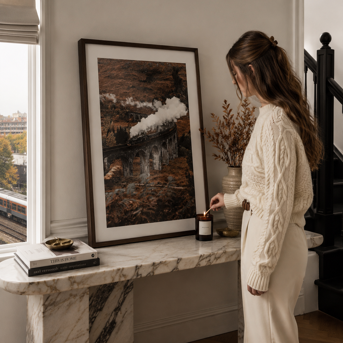 A woman in a cream cable-knit sweater and matching trousers stands beside a marble console table, looking at a large framed landscape art print leaning against a wall.