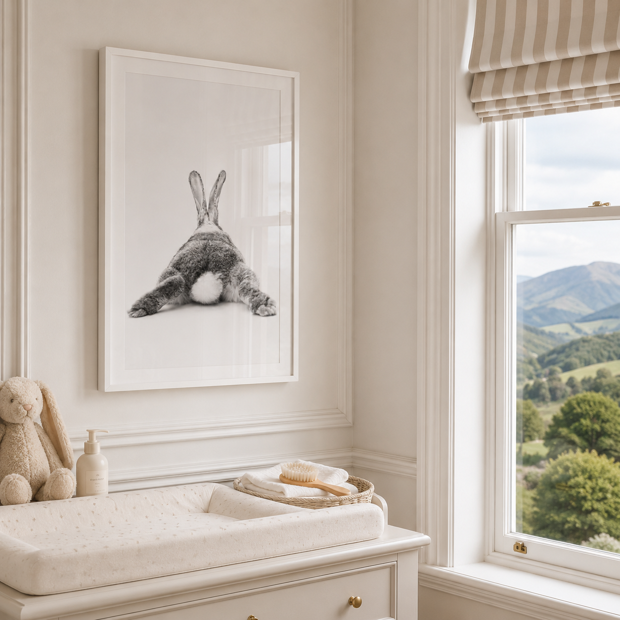 A framed black and white art print of a rabbit sitting in a baby nursery, hanging on a cream wall above a changing table with toys and a window showing a green mountain landscape.