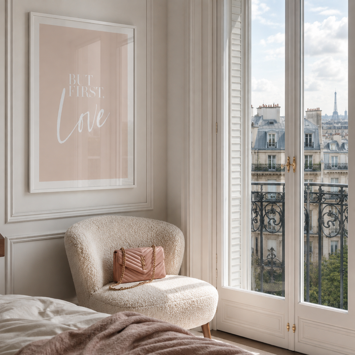 A framed wall art print with the words 'But First, Love' in white script on a soft blush background, hanging above a cream bouclé armchair with a pink quilted handbag in a bright Parisian-style bedroom. Large white French doors with a balcony railing and distant Eiffel Tower view are visible.