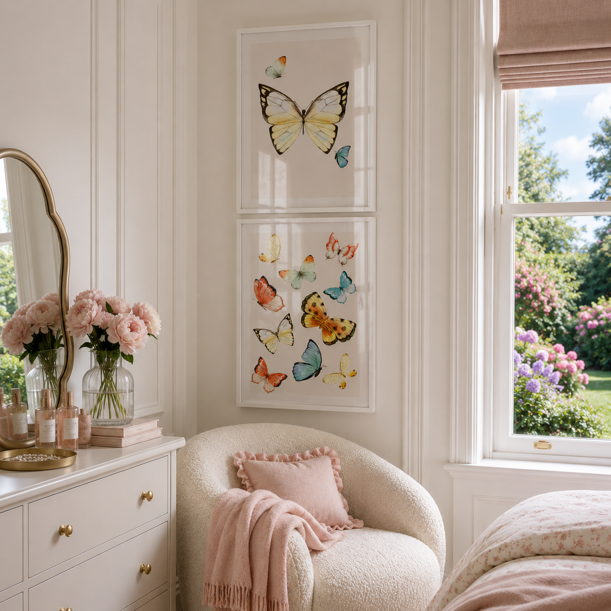 A styled bedroom corner with two vertically stacked framed butterfly art prints on a cream wall above a blush pink armchair. A white dresser with gold handles, a mirror, and pink flowers are visible, with a window showing a garden view to the right.