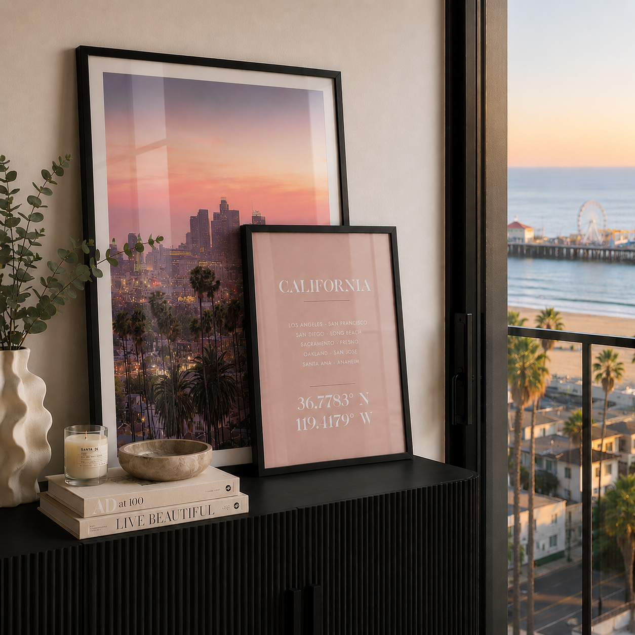 Two framed art prints displayed on a black console table in a modern room setting, with a window showing an ocean view. The larger print features a colorful sunset city skyline, and the smaller print has a pink background with white text reading 'CALIFORNIA' and coordinates.