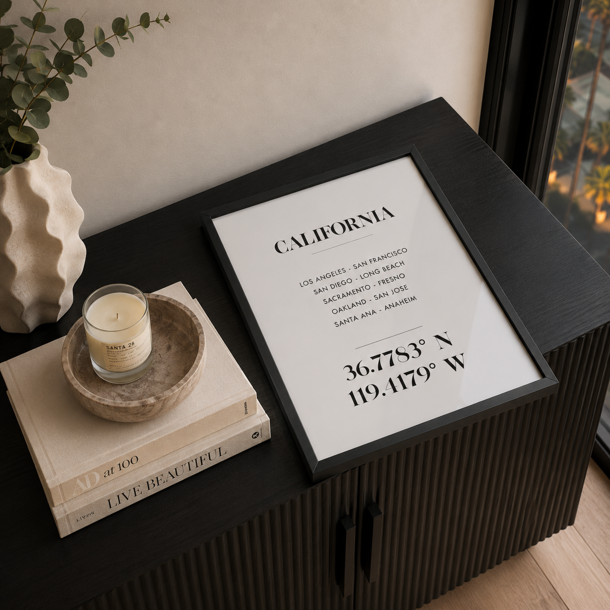 A framed California city list print with coordinates, styled on a dark ribbed cabinet beside a candle, books, and a vase in an indoor setting.