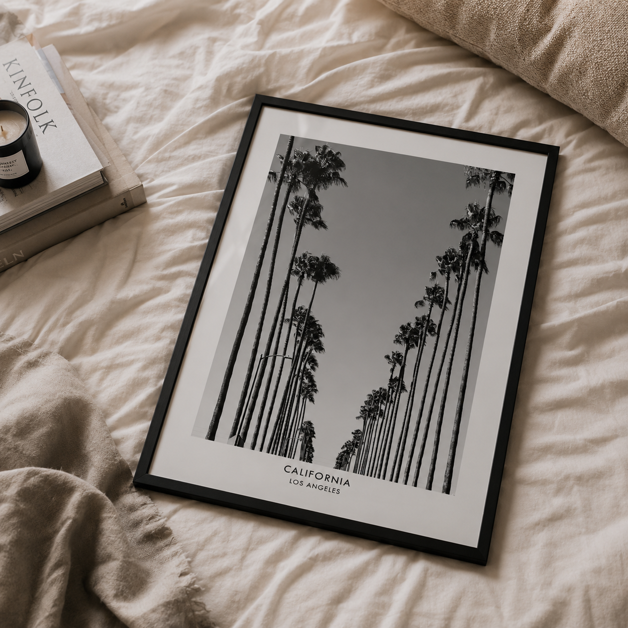 A framed black and white art print featuring tall California palm trees, styled on a beige linen bed next to a book and candle.
