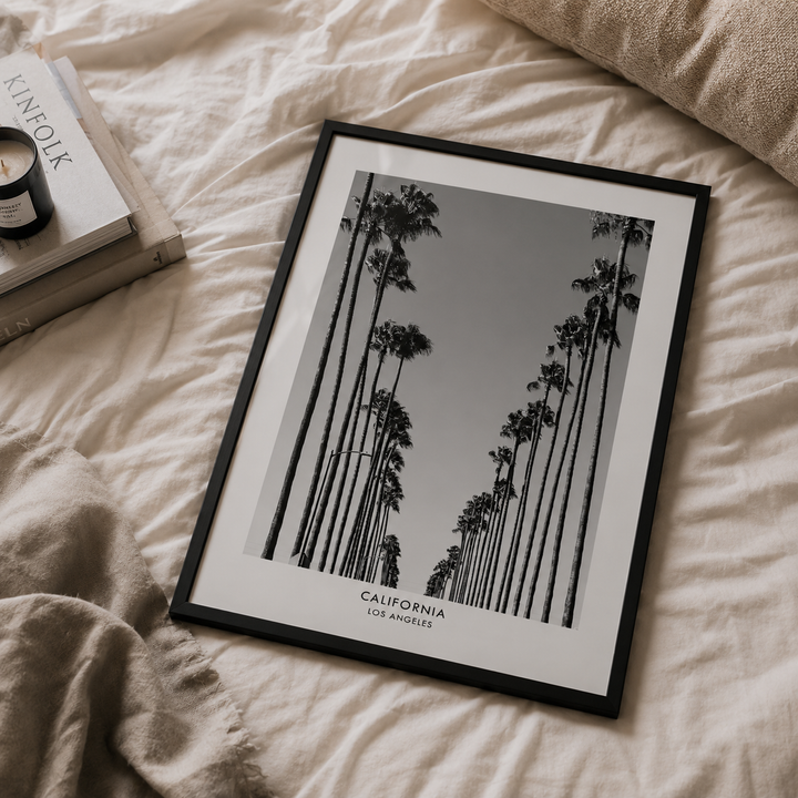 A framed black and white art print featuring tall California palm trees, styled on a beige linen bed next to a book and candle.