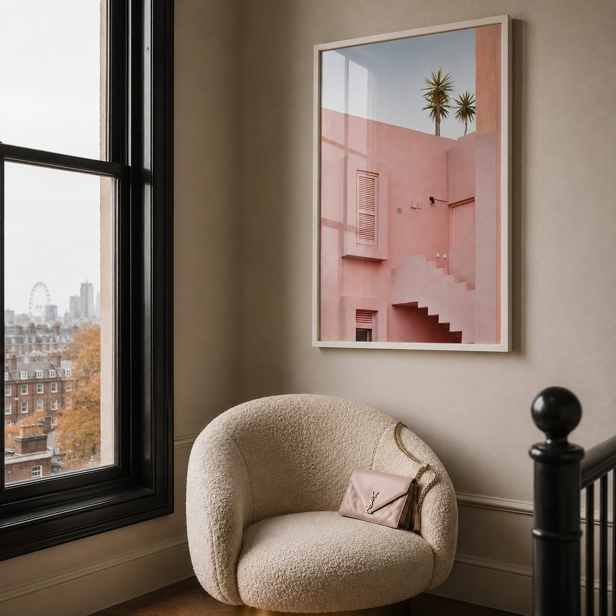 A framed art print featuring a pink modernist building with stairs and palm trees, displayed on a beige wall in a stylish room above a cream bouclé armchair.