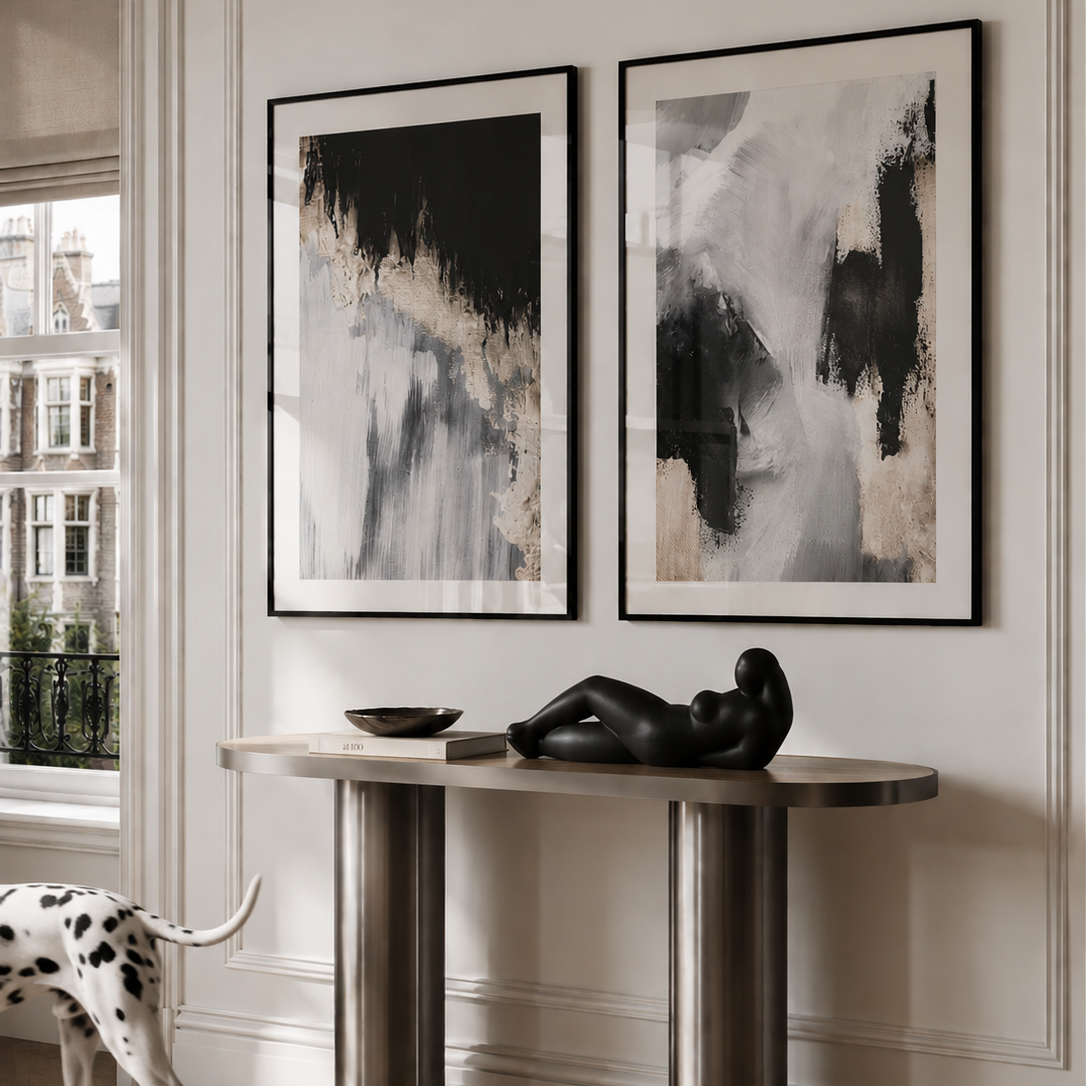 Two framed abstract black, white, and beige wall art prints hang above a modern console table in a bright interior. A black sculptural figure sits on the table, and a white dog with black spots walks in from the left near a window.