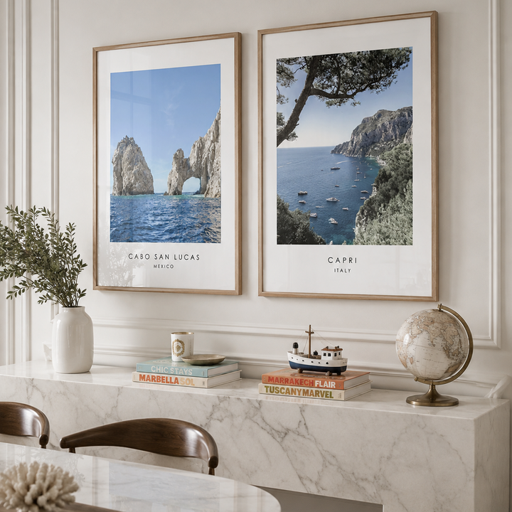 Two framed travel art prints of Capri and Cabo San Lucas hang on a cream paneled wall above a marble console table in a styled dining room.
