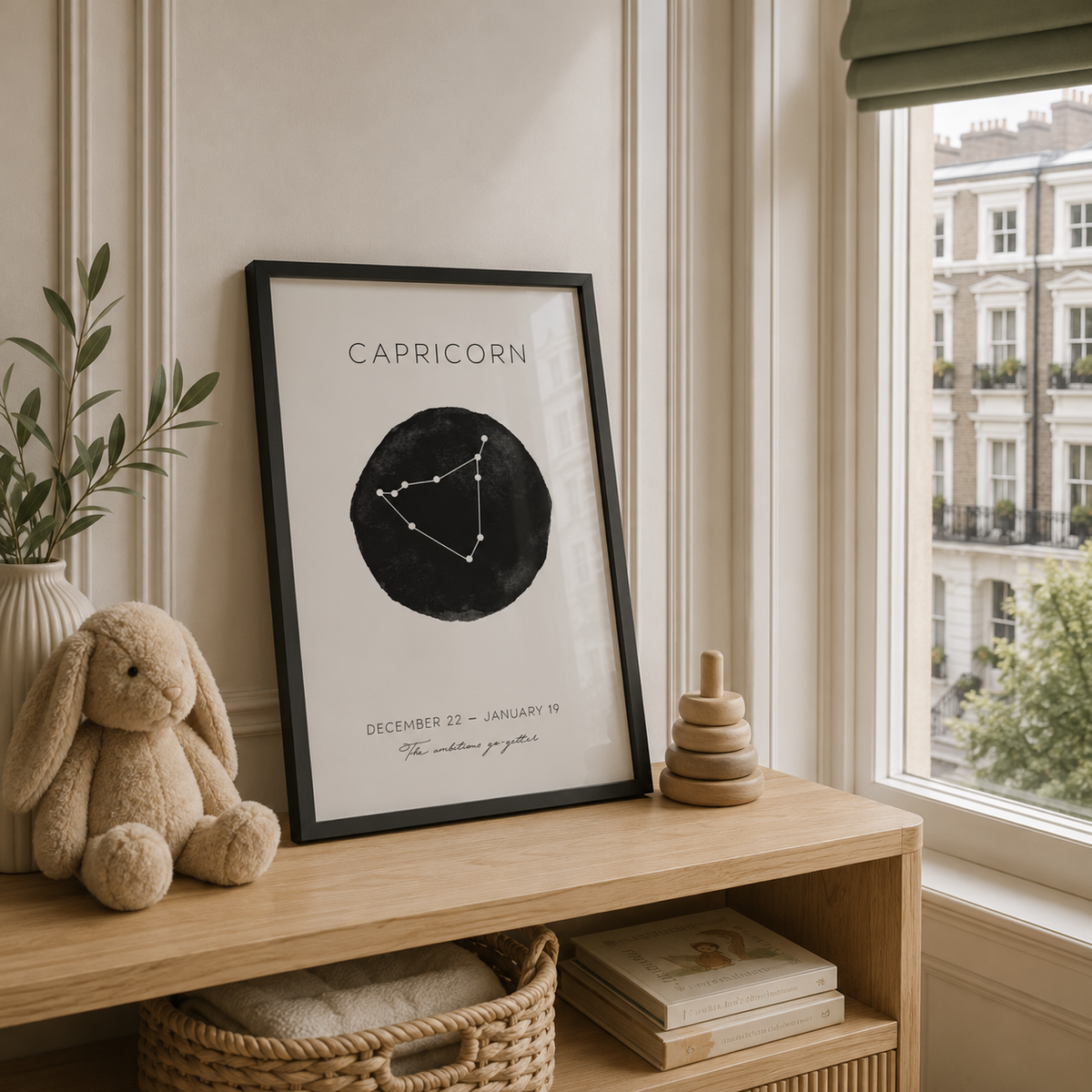 A framed Capricorn art print with a black circular constellation graphic and elegant text, leaning on a light wood console table in a softly styled room with a plush bunny, basket, and window view.