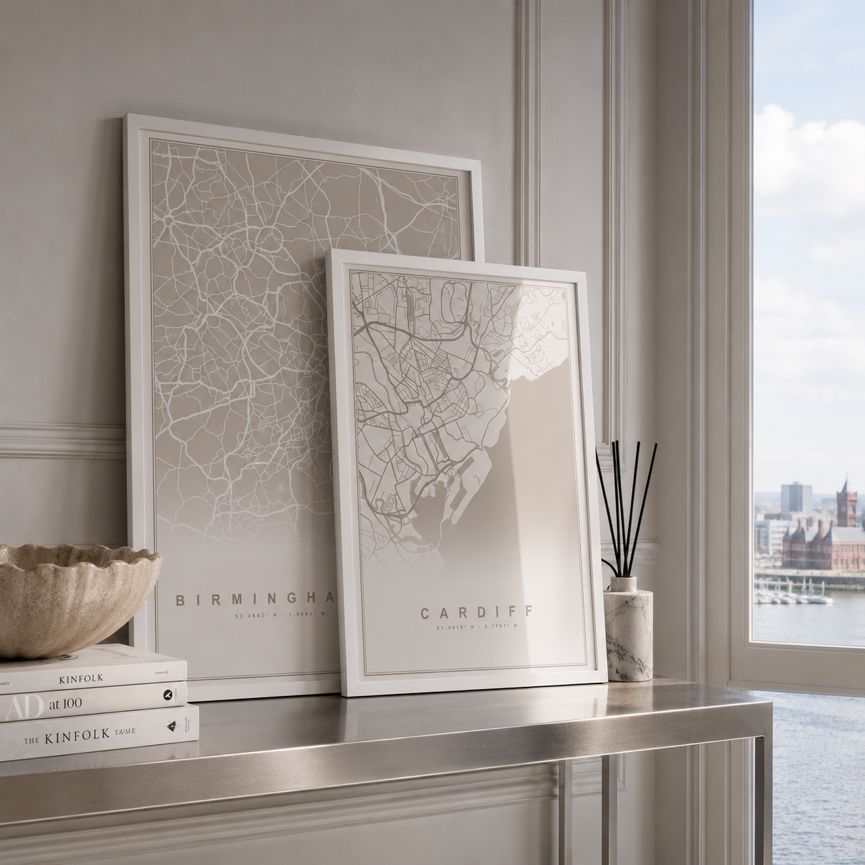 Two framed city map art prints leaning on a console table in a bright interior. The larger print reads Birmingham and the smaller print reads Cardiff.
