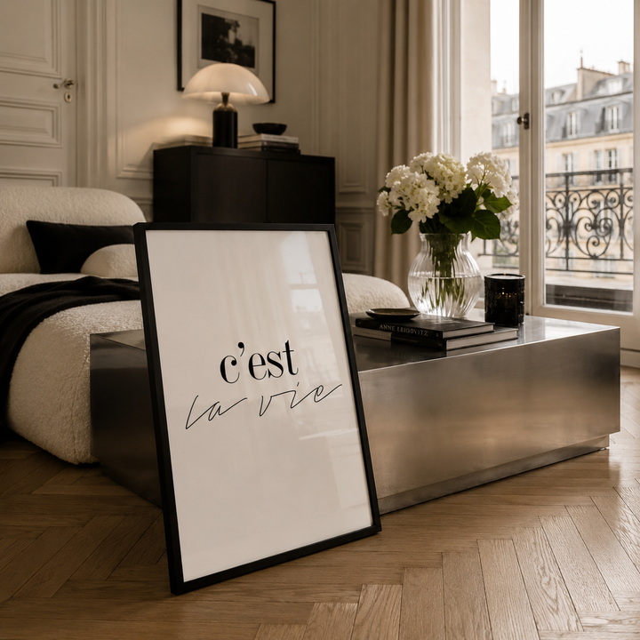 A framed French wall art print leaning against a modern coffee table in a stylishly decorated bedroom. The print features the text 'c'est la vie' in elegant black script on a white background with a thin black frame.