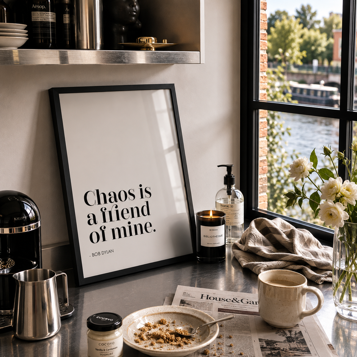 A framed typographic art print with the quote 'Chaos is a friend of mine. - Bob Dylan' leaning on a kitchen counter beside a window, with a mug, plate, candle, and flowers nearby.