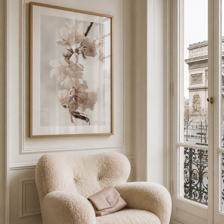 A large framed botanical art print of pale pink blossoms hangs on a cream paneled wall above a plush cream bouclé armchair with a matching YSL clutch bag, next to a window overlooking a Parisian building.