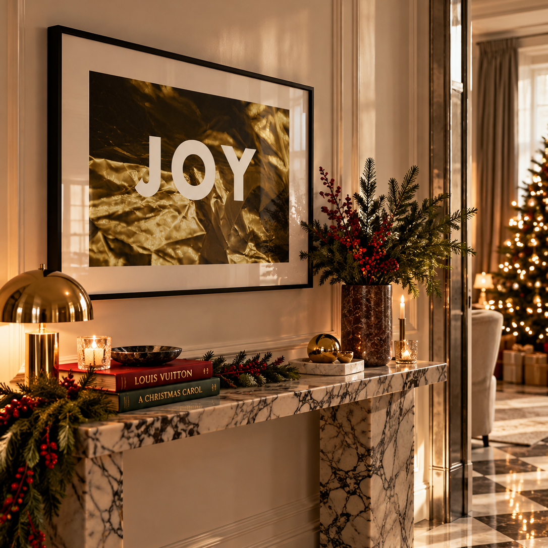 A framed wall art print with the word 'JOY' in white over a dark textured background, displayed above a marble console table in a decorated holiday interior. The scene includes candles, greenery, red berries, and a Christmas tree in the background.