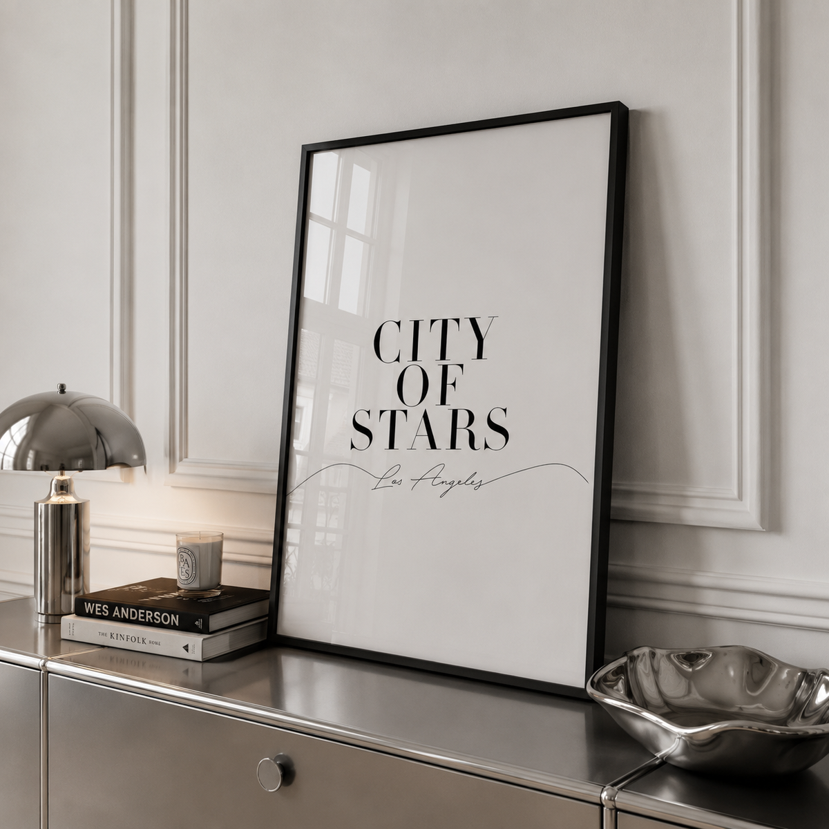 A framed typographic wall art print reading 'CITY OF STARS' with 'Los Angeles' in script, leaning on a metallic console table against a white paneled wall. Decorative objects including a lamp, books, and a bowl are visible in the room setting.