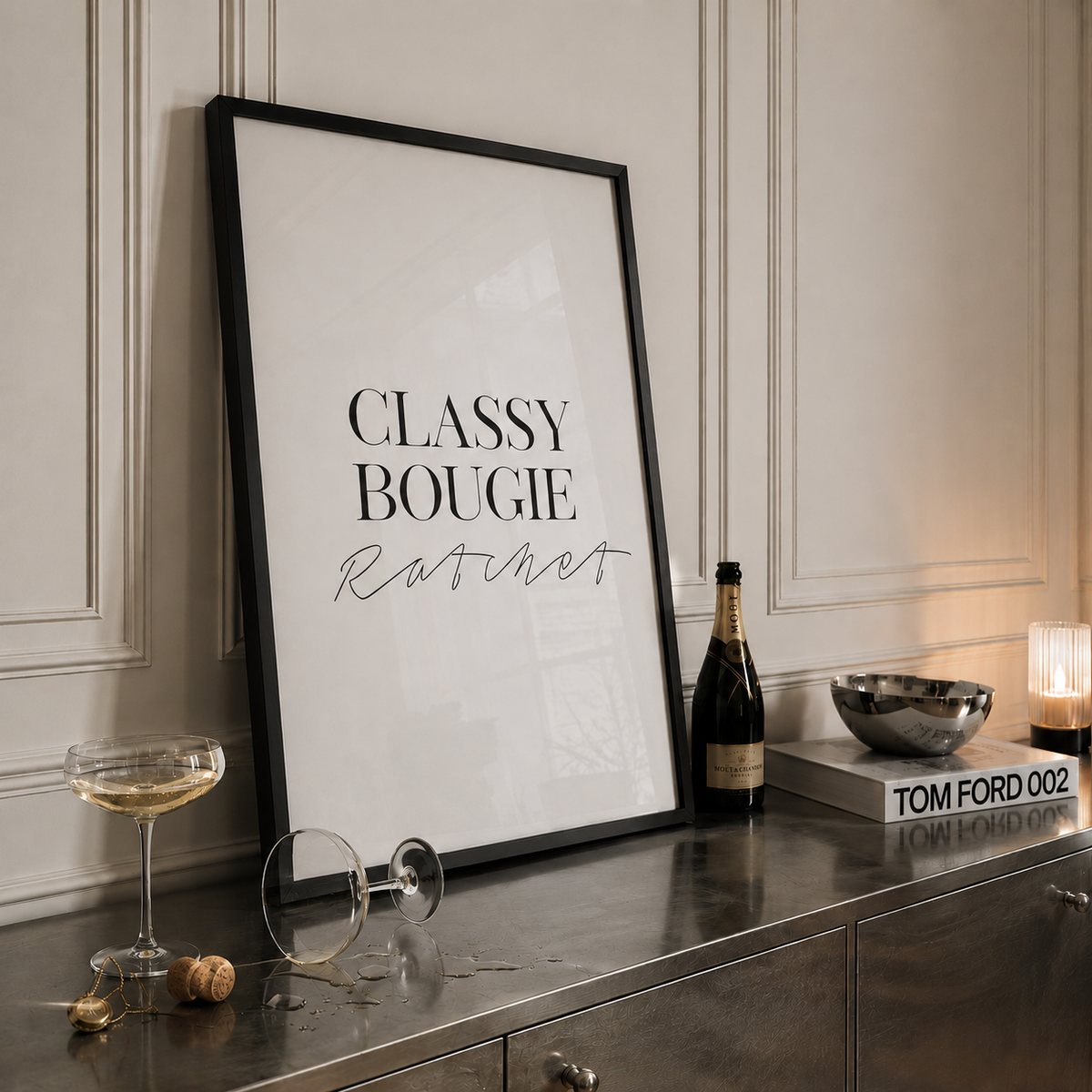 A framed typographic art print leaning against a paneled wall on a metallic console table. The print has a black frame and reads 'CLASSY BOUGIE Ratbert' in black lettering on a white background.