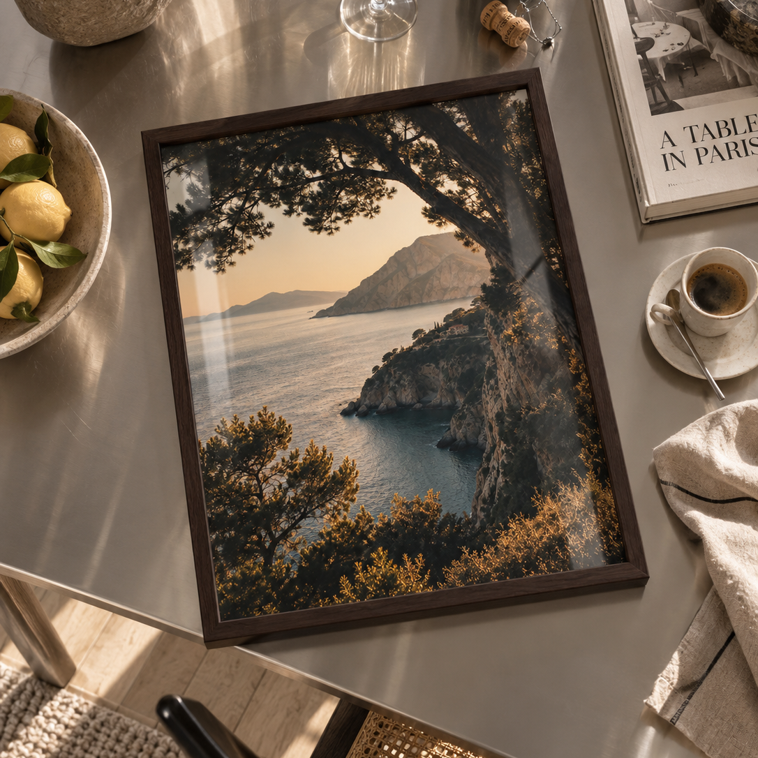 A framed coastal landscape print displayed on a metallic table, surrounded by dining and lifestyle props including a wine glass, book, coffee cup, and lemon bowl.