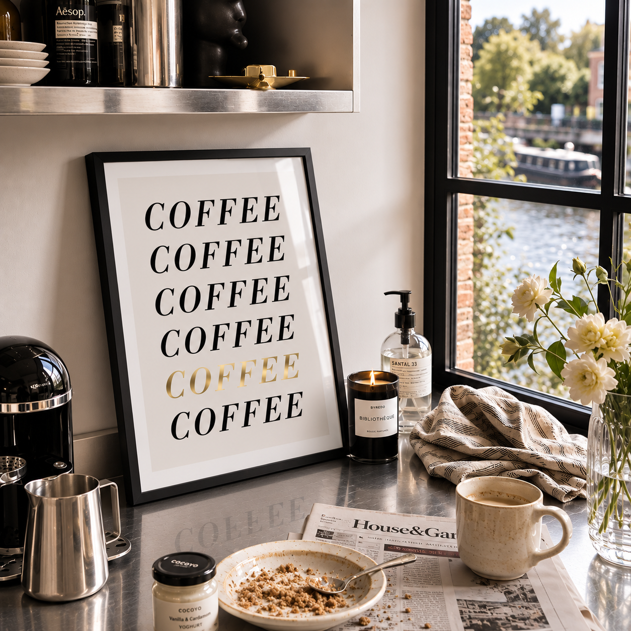A framed coffee-themed wall art print with the word 'COFFEE' repeated in black and one word in gold foil, leaning on a kitchen counter beside a mug, newspaper, candle, and flowers by a window.
