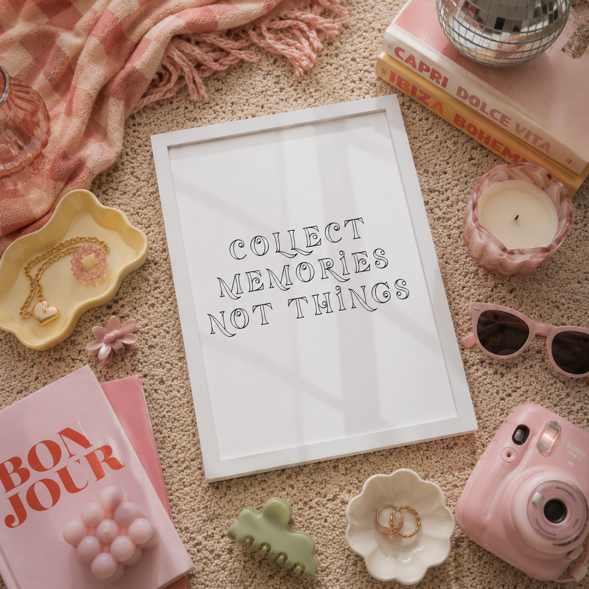 A white framed typographic art print reading 'COLLECT MEMORIES NOT THINGS' styled in a pink flat-lay with a pink camera, sunglasses, candle, books, jewelry dish, and scarf on a beige carpet background.