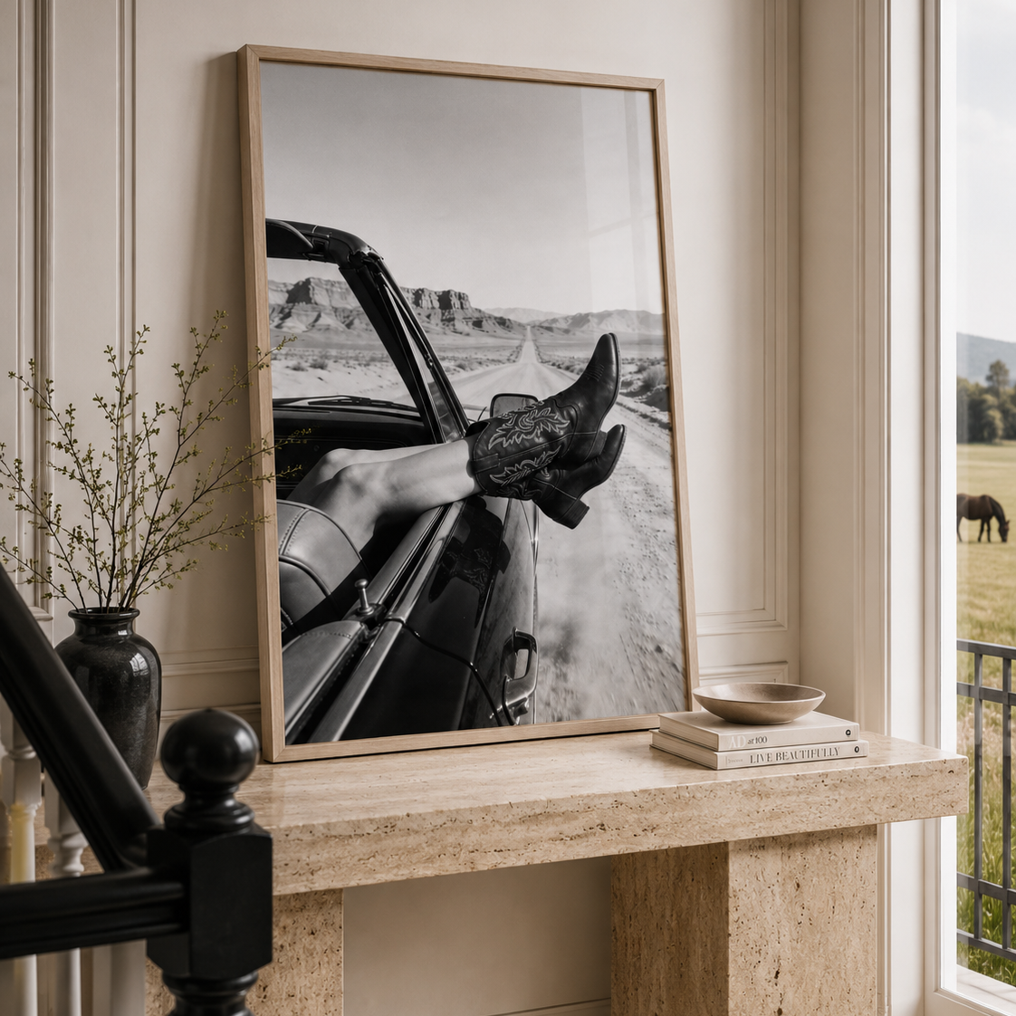 A framed black and white photograph leaning on a stone console table in a bright room. The print shows a person in a convertible car with boots, and the room includes a window, vase with dried stems, and a small bowl.