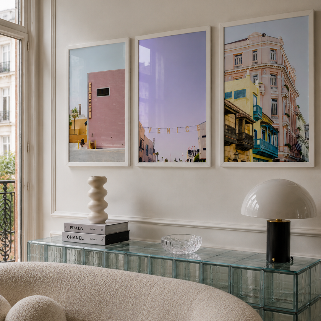 Three framed vertical art prints depicting colorful Venice scenes hang on a white wall above a glass side table in a modern living room setting.