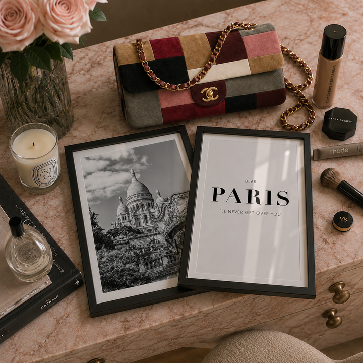 Two framed wall art prints displayed on a pink marble vanity table, surrounded by lifestyle props including a patchwork Chanel bag, a candle, perfume, and cosmetics. The left print is a black-and-white photograph of the Sacre-Coeur de Paris, and the right print features the text 'DEAR PARIS I'LL NEVER GET OVER YOU' in black on a white background.