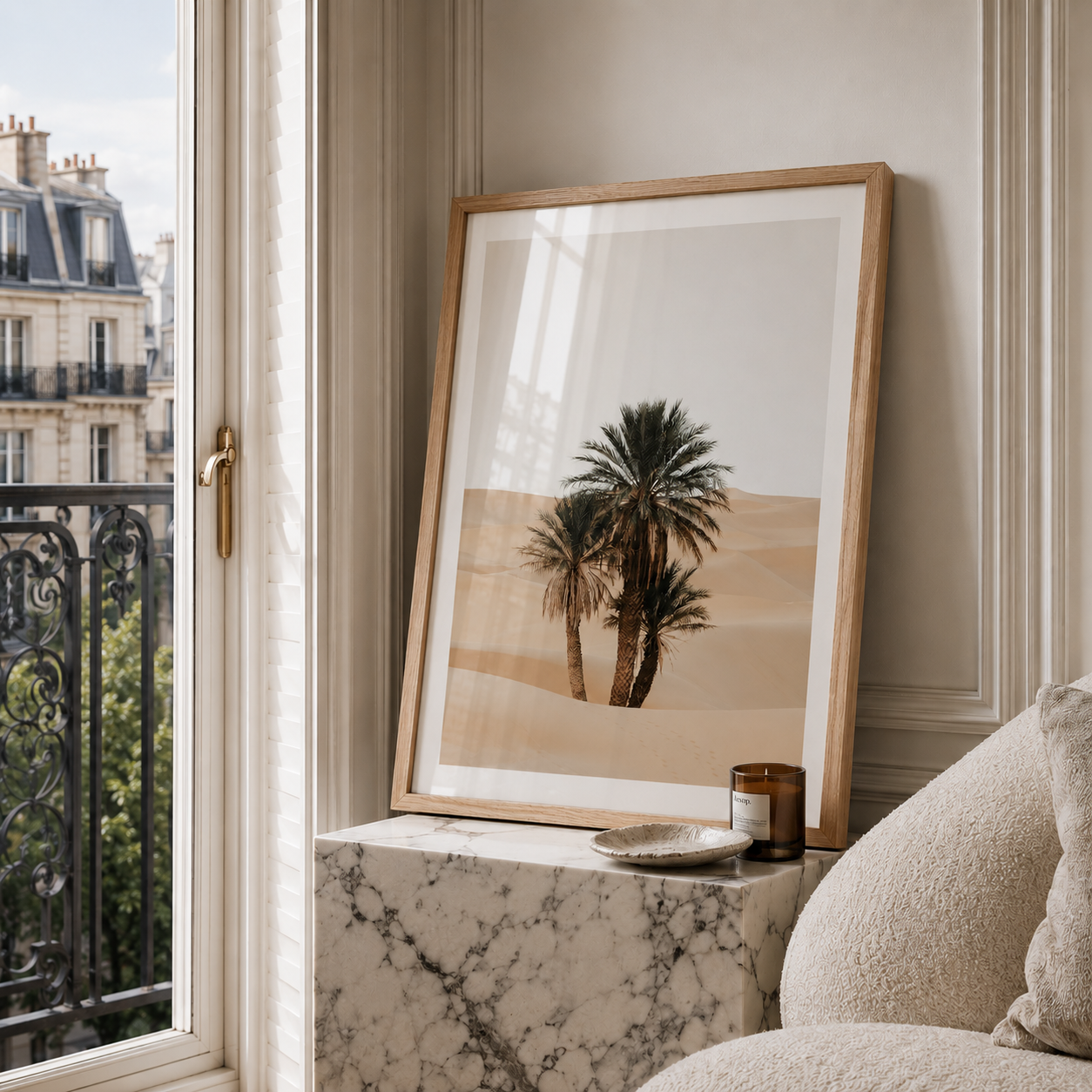 A framed art print featuring two palm trees in a desert landscape, leaning on a marble side table next to a window with a view of Paris buildings.