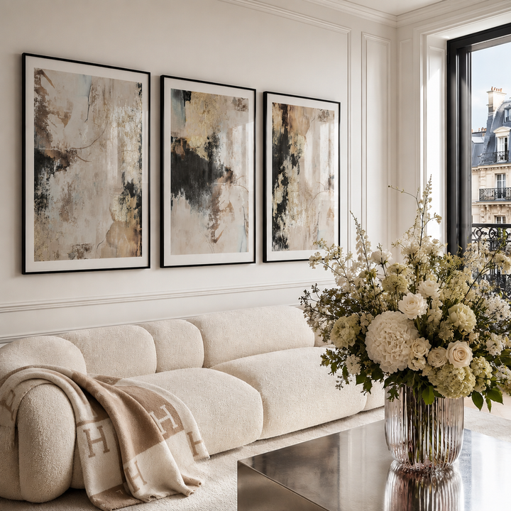 Three framed abstract wall art prints in black frames hang above a cream boucle sofa in a bright living room. A large vase of white and cream flowers sits on a reflective table in the foreground, and a window on the right shows a Parisian-style building exterior.