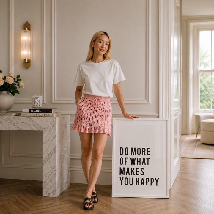 A woman stands in a bright, elegant room wearing a white short-sleeve t-shirt and a pink pleated crochet mini skirt. She is leaning against a white sign that reads 'DO MORE OF WHAT MAKES YOU HAPPY'.