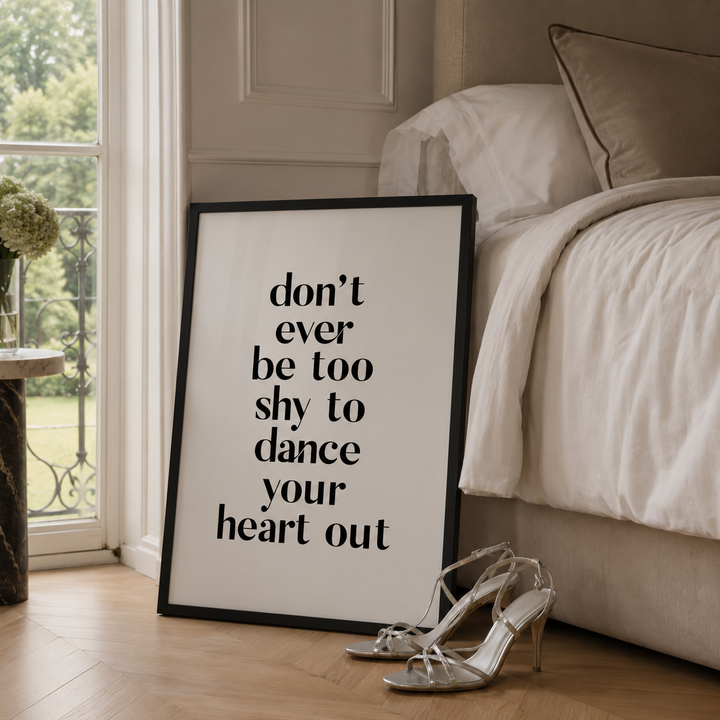 A framed typographic wall art print leaning against a bed in a softly lit bedroom. The print has black text reading “don’t ever be too shy to dance your heart out” on a white background with a black frame.
