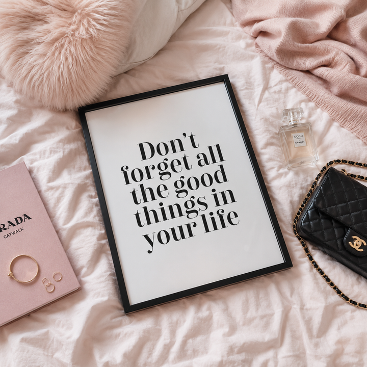 A framed typographic wall art print with the quote 'Don't forget all the good things in your life' in black serif text on a white background, styled on a blush pink bed with a pink pillow, pink blanket, a black quilted Chanel handbag, a pink book, and small jewelry accessories.