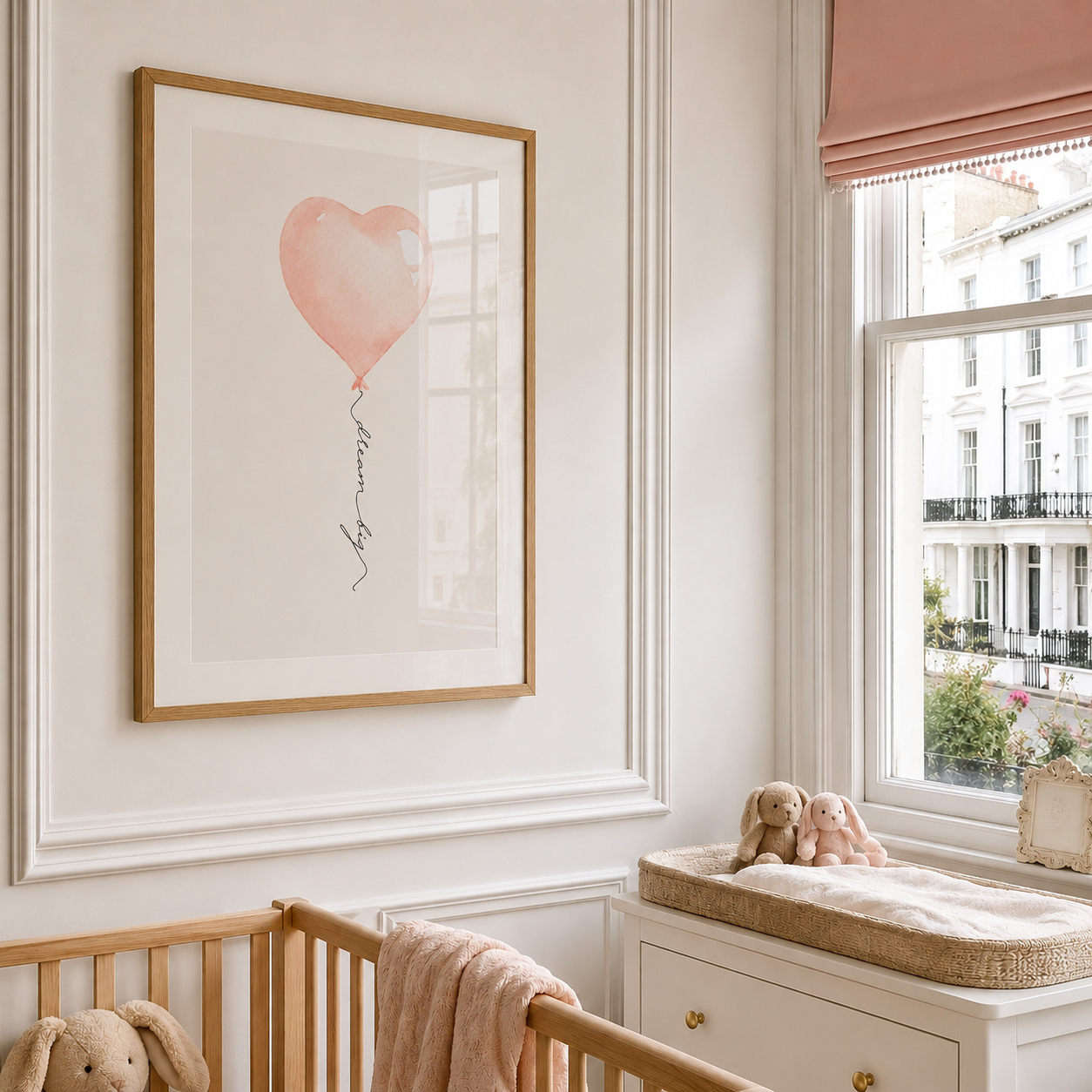 A framed art print featuring a soft pink heart-shaped balloon with the word 'Love' written below, displayed on a nursery wall above a wooden crib. The room has white paneling, a window with a pink blind, and a changing table with stuffed toys.