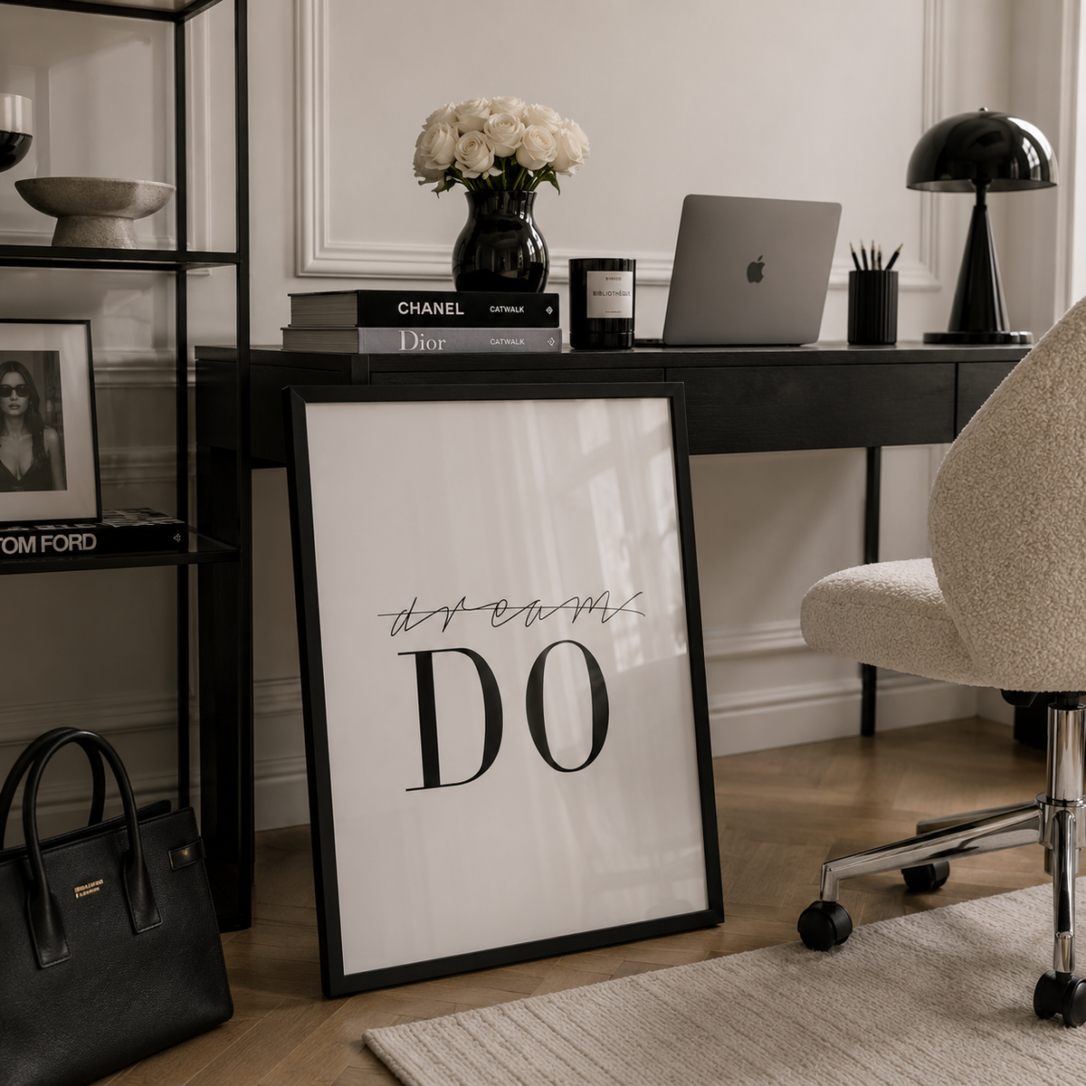 A framed motivational art print leaning against a desk in a modern home office setting. The print features the text 'dream DO' in black typography on a white background, enclosed in a black frame.