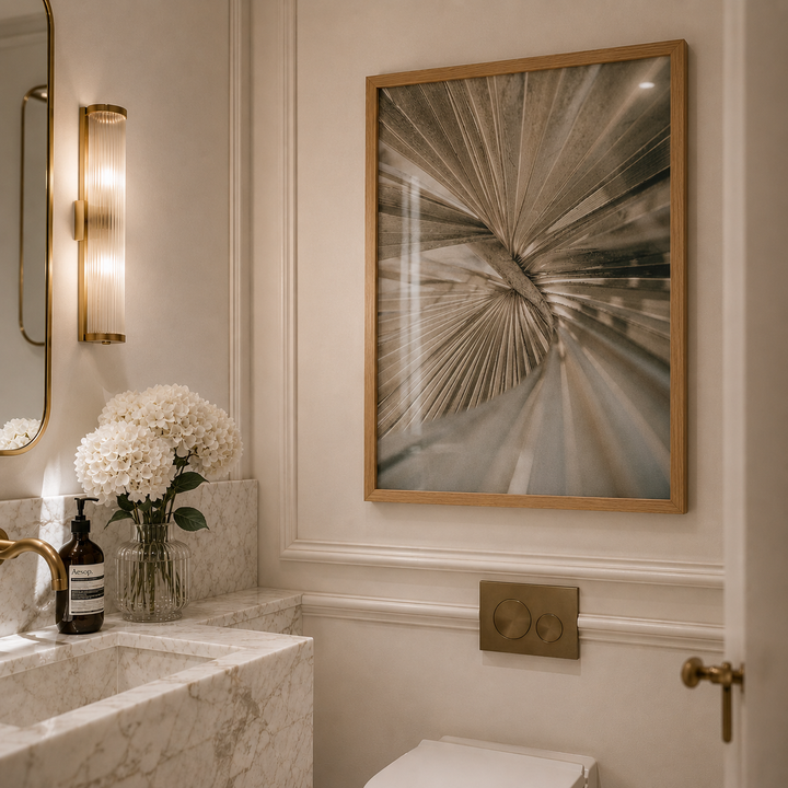 A framed abstract art print featuring a radial burst pattern in warm beige, brown, and gray tones, hanging on a cream-colored wall in a modern bathroom setting.