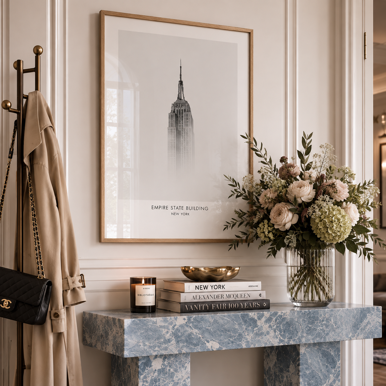 A framed black and white art print of the Empire State Building hangs on a cream paneled wall above a blue marble console table styled with a book stack, candle, gold bowl, and a vase of blush and white flowers.